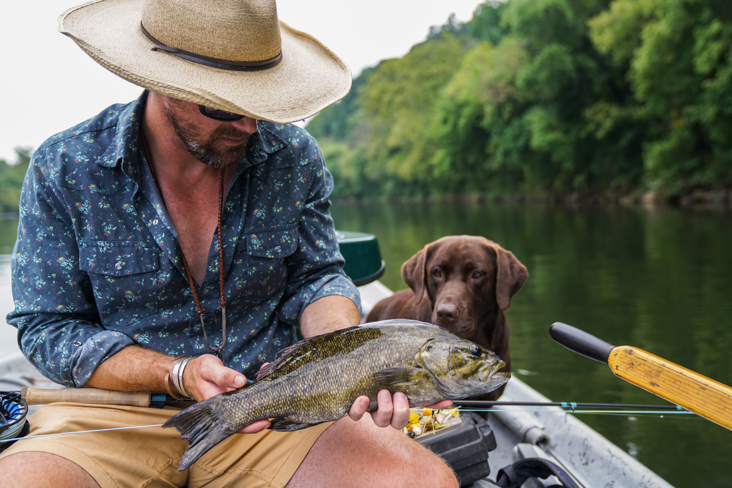 Man in a wide-brimmed hat holding a smallmouth bass in a boat, with a brown dog looking on and a forested riverbank in the background.