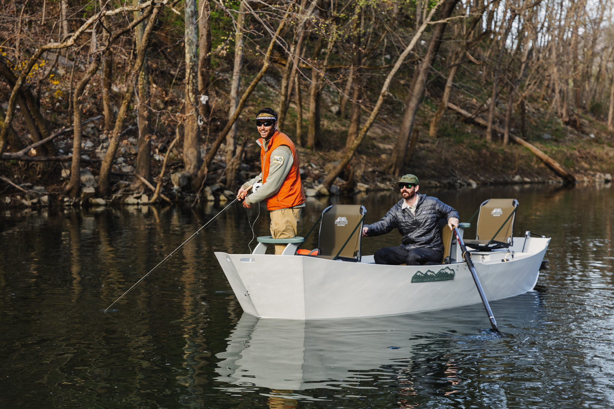 Two men in a Blue Ridge drift boat fly fishing on a calm Virginia river surrounded by bare trees in early spring or late fall.