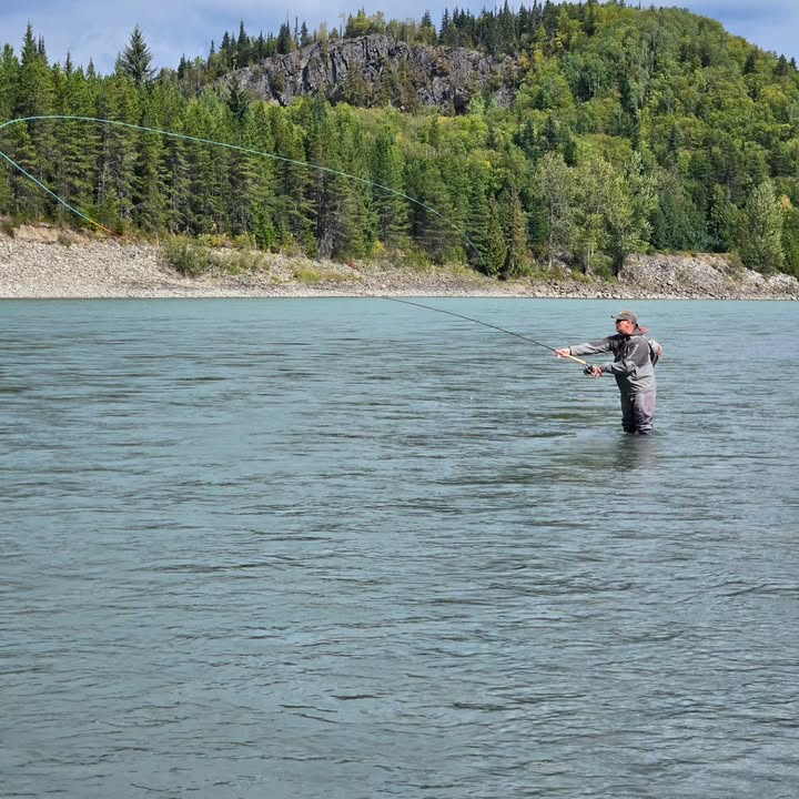 Calvin Tull casting his line in a serene river, surrounded by lush forested hills in the background, showcasing the art of fly fishing in a peaceful natural setting