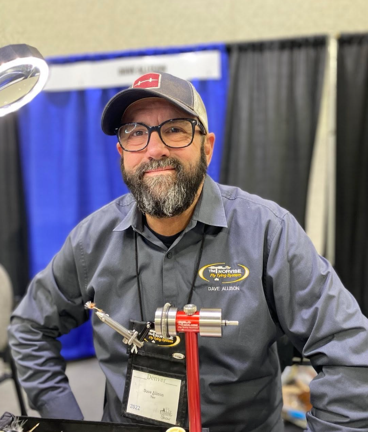 Dave Allison tying a dry fly on a Nor-Vise at a fly tying expo, sitting at a tying station with materials and tools in front of him.