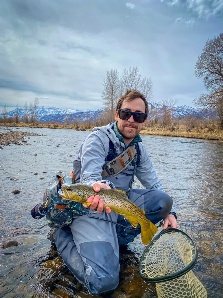 Pablo Signori holding a brown trout while fly fishing in a river with mountains in the background
